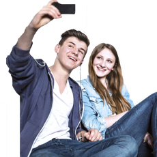 Two young people, a boy and a girl, are taking a photo with a mobile phone. The boy is turned away and holds the device while the girl smiles facing forward.