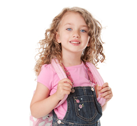 Girl with rosy cheeks, curly hair and a backpack with drawings, wearing denim overalls and a pink shirt.