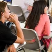 Image of several students in a classroom, sitting in chairs with backs, with one student handing water to another.