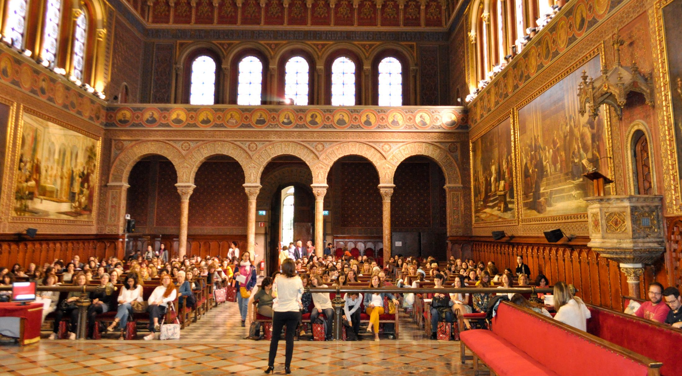 Wide space with brick walls, arched windows and tiled floor. People are seated in rows. Decorative elements are on both sides of the room.