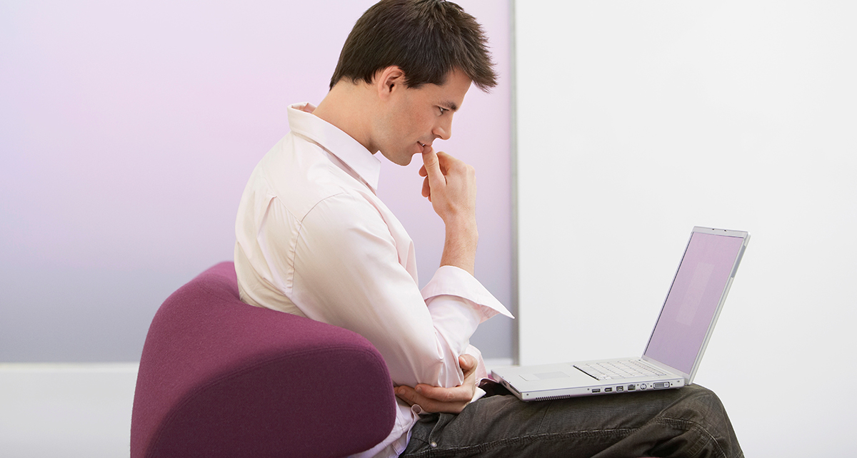 A young man with dark hair and a buttoned shirt, sitting in a purple chair in front of a laptop. He is touching his eyebrow with his hand and appears to be concentrating on the computer.