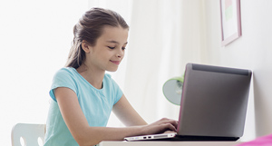 Una niña adolescente, sentada frente a un portátil, escribe en el teclado. Tiene el pelo recogido y viste una camiseta azul claro. A su lado, se ve una pared con un cuadro colgado.