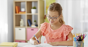 Niña con gafas escribiendo en un libro de texto, con un lápiz y un fondo de útiles escolares.
