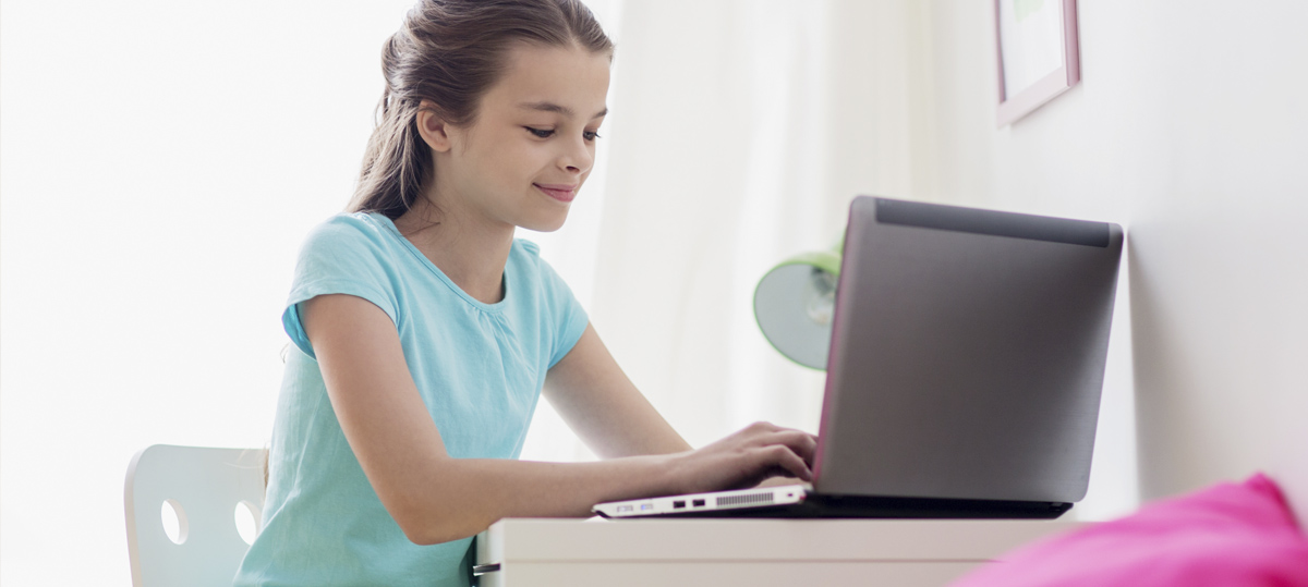 Una niña adolescente, sentada frente a un portátil, escribe en el teclado. La imagen muestra un espacio de estudio con un escritorio y un cojín de color rosa.