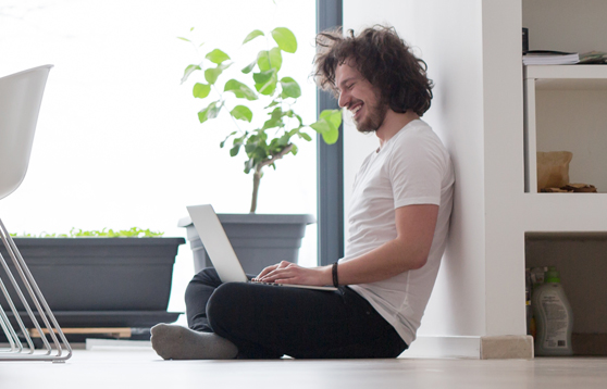 Un hombre joven, sentado sobre el suelo de una habitación, usando una laptop. Tiene cabello oscuro y un gesto de alegría al leer el contenido de la pantalla. Un pequeño ejemplar de planta verde se encuentra al fondo.