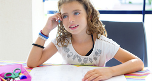 Niña con gafas y cabello ondulado, sentada frente a un escritorio con material de dibujo. Tiene una mano apoyada en el escritorio y otra sosteniendo un lápiz.