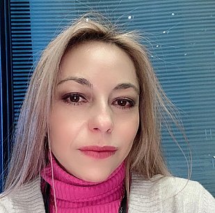 Retrato de una mujer de mediana edad con cabello castaño y ojos marrones, vestida con un suéter rosa y una chaqueta. Tiene auriculares y una expresión seria.