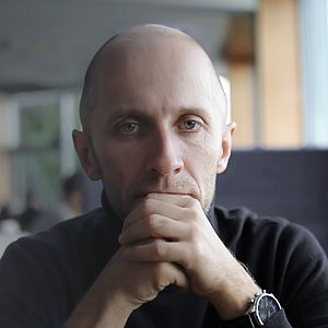 Retrato de hombre adulto, de rostro redondo y cabello calvo, con expresión seria y ceño fruncido. Tiene la mano derecha sobre la boca y la izquierda entreabierta. Viste una camisa negra y lleva un reloj en la muñeca.
