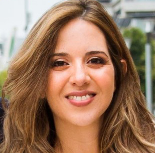 Fotografía de una mujer con cabello castaño ondulado y maquillaje. Tiene una sonrisa mostrando sus dientes y una expresión amable.